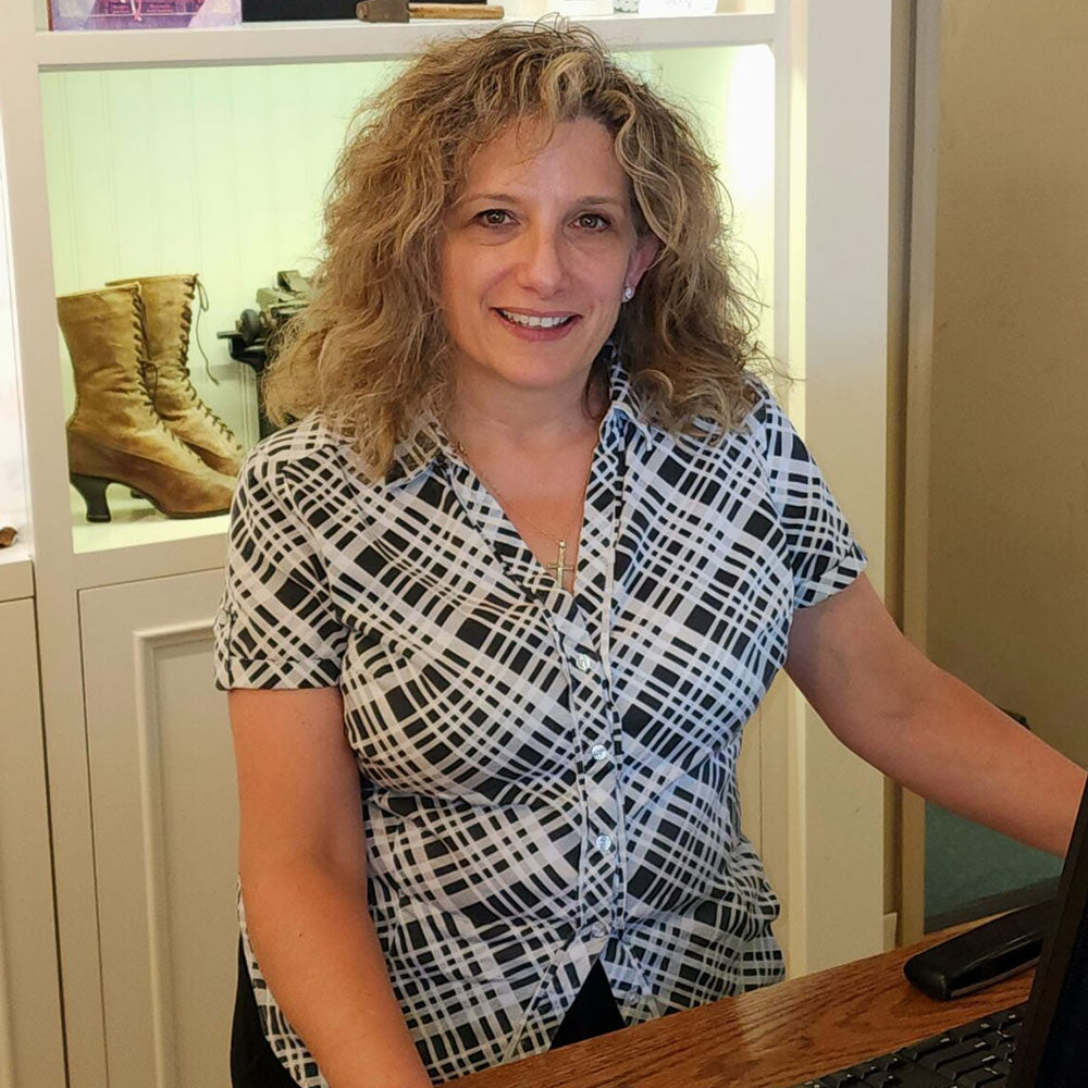 Michele in a patterned shirt sitting at a desk with shoes displayed in the background