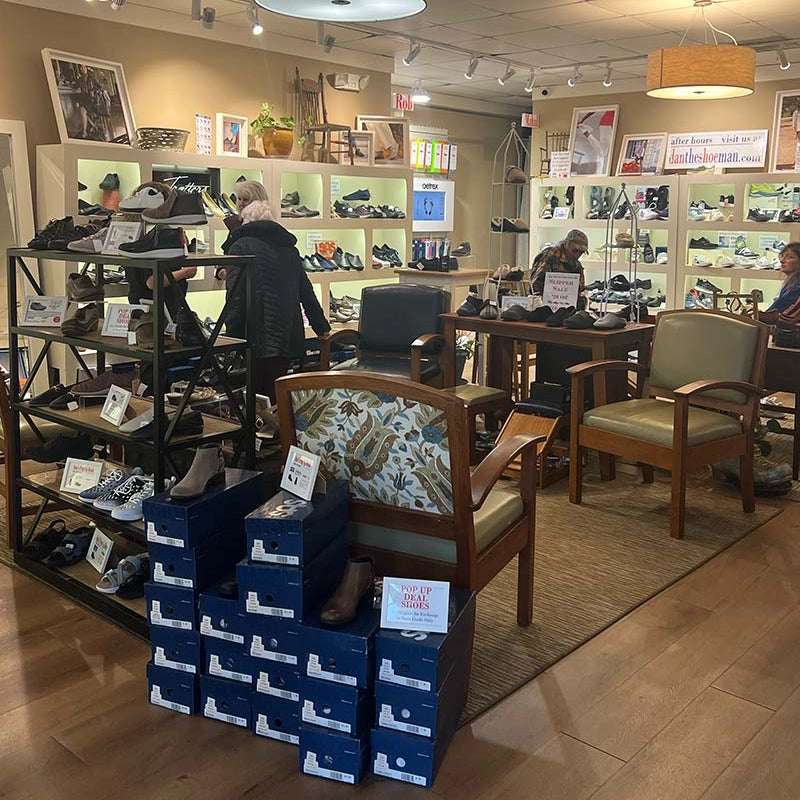Shoe store interior with shoe boxes, chairs, and people browsing shoes.